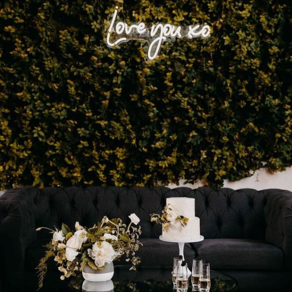 Love You XO LED neon sign on a green photo wall with a floral arrangement and cake in the foreground  - sign by CustomNeon.co.uk / photo by candileonardphotography.com