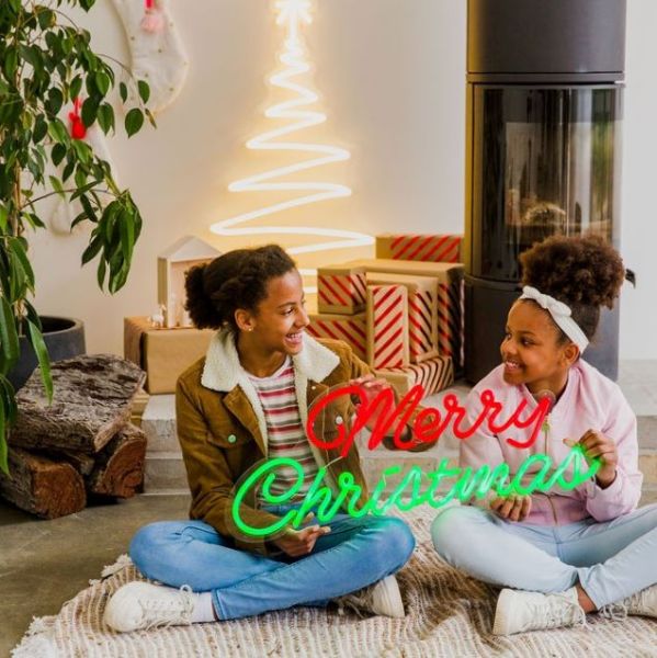 Merry Christmas Red & Green LED Neon Sign by CUSTOM NEON® held by smiling girls with a Nativity scene, presents & decorations in the background.