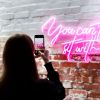 Person taking a photo of a pink LED neon sign reading You Can't Sit with Us - Custom Neon