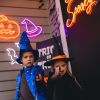 Custom Neon® Halloween lightup signs as door & porch decorations: jack-o-lantern, purple witch's hat, bats & Stay Spooky quote sign with kids in costumes pointing wands