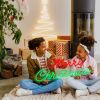 Merry Christmas Red & Green LED Neon Sign by CUSTOM NEON® held by smiling girls with a Nativity scene, presents & decorations in the background.