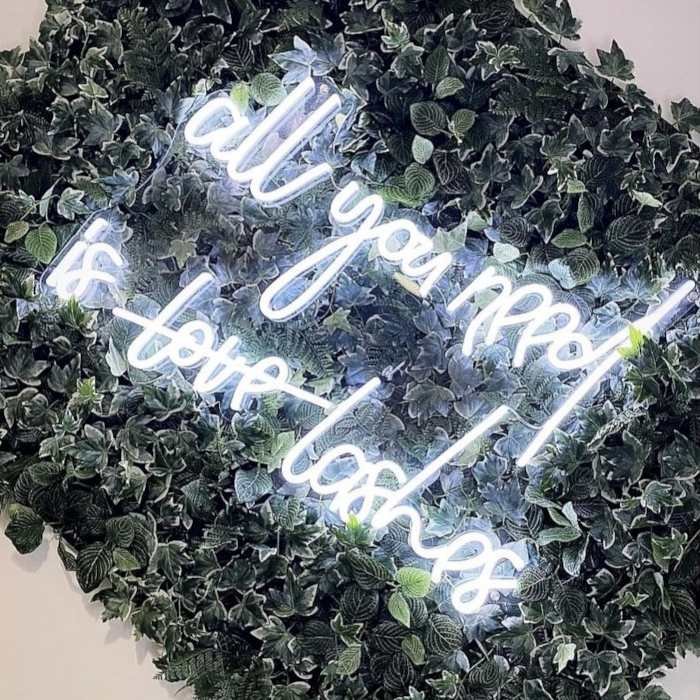 All You Need is Lashes white Custom Neon® sign on a greenery wall in a lash bar