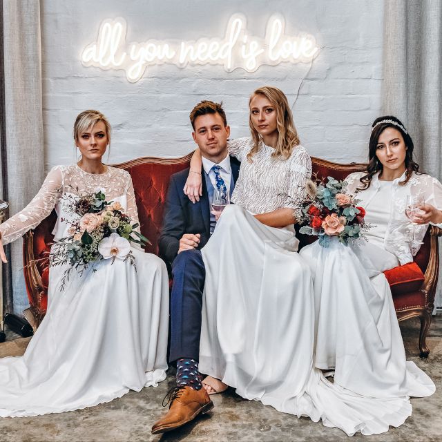 White All You Need is Love Custom Neon® sign on a white brick wall behind the bridal party sitting on a red velvet couch