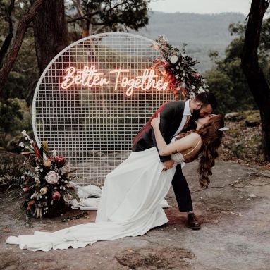 Custom Neon® red Better Together sign on a circular mesh frame outdoors behind the bride and groom embracing