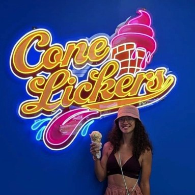 Custom Neon® colorful logo sign on a dark blue feature wall behind a customer taking a selfie @conelickers