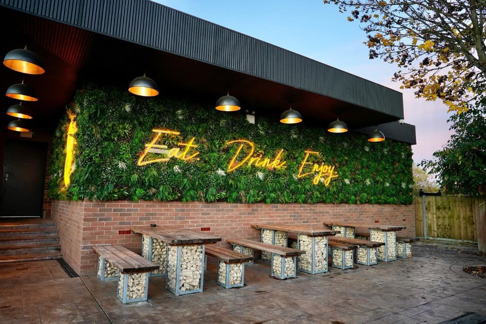 Yellow Custom Neon® signs of the Copper Fox Bar and Grill circular logo and the words Eat Drink Enjoy mounted on a lush greenery wall above dining tables in a pub garden