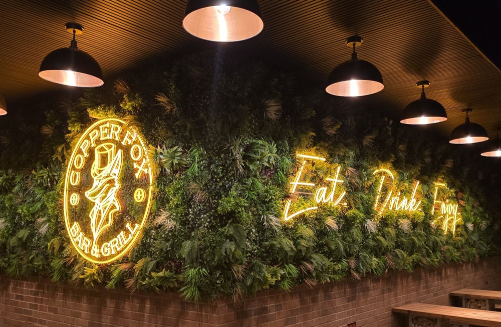 Yellow Custom Neon® signs of the Copper Fox Bar and Grill circular logo and the words Eat Drink Enjoy mounted on a lush greenery wall above dining tables in a pub garden