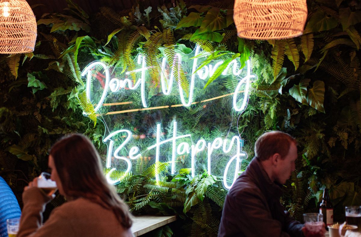 Don't Worry Be Happy Custom Neon® sign on greenwall with diners in the foreground @mktplce.uk photography @brianfbenton