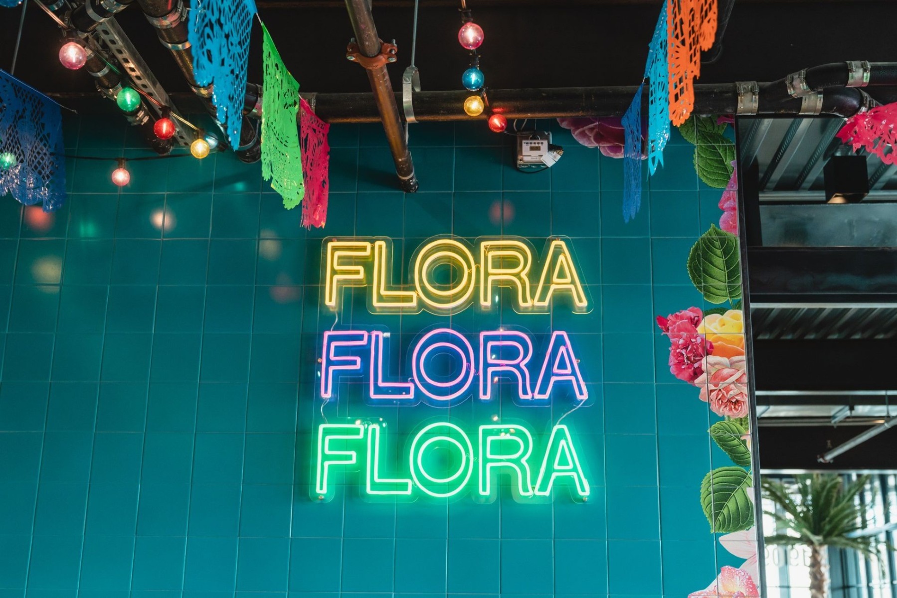 Yellow blue and green Custom Neon® restaurant name sign on a blue tiled wall with a mural border @florarestaurantnm @heritagerec