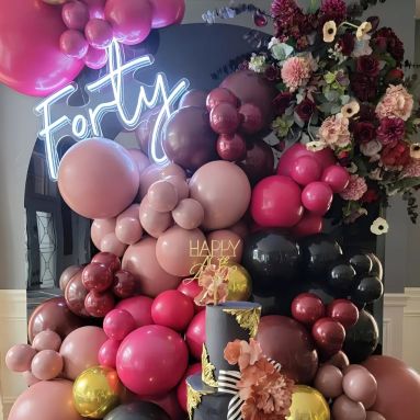 Custom Neon® Forty milestone birthday sign on a glossy black arch surrounded by deep red and pale pink flowers and a maroon and black balloon garland - event decor by @decorandfiesta