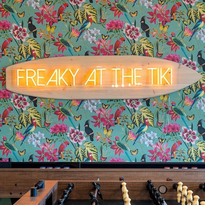 Freaky At The Tikki orange LED Custom Neon® sign on a small wooden surfboard against tropical wallpaper above a fuse ball table - interior design by Somerled