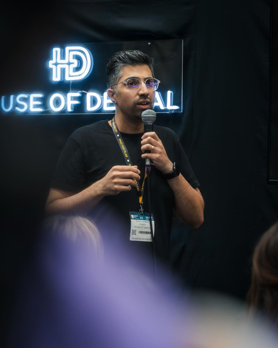 Man speaking at a conference with a black backdrop featuring a white House of Dental Custom Neon® sign