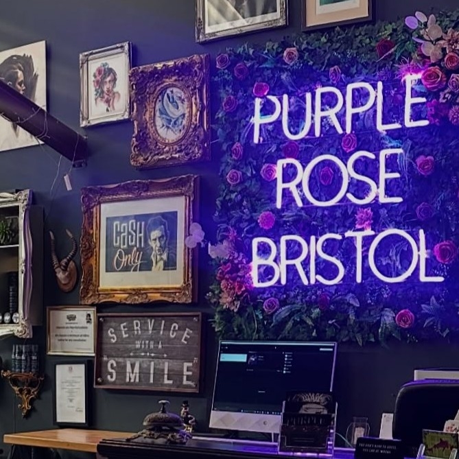 Purple Custom Neon® sign reading Purple Rose Bristol mounted on a rose wall behind the front desk @purplerosetattoo