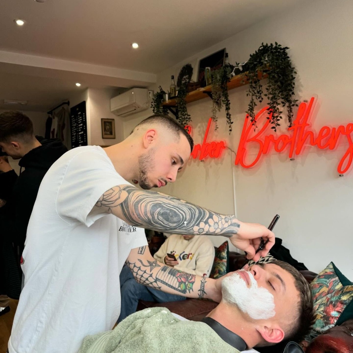 The Barber Brothers red Custom Neon® sign on the wall behind a barber shaving a client